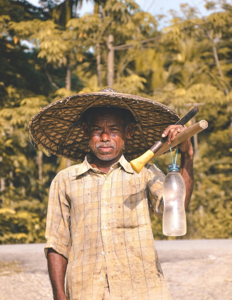 An Indian farmer holding tools and a bottle in a rural setting.